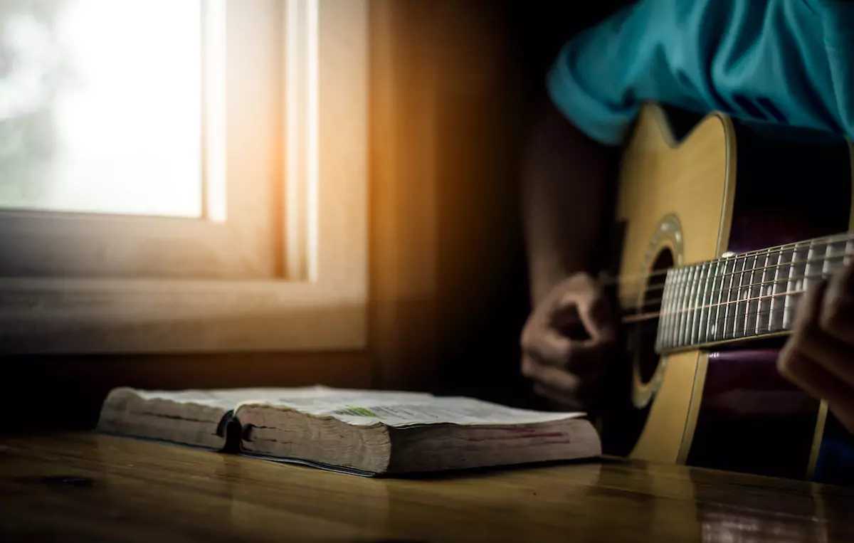 Young man is playing guitar and sing a song from Christian hymn book with bible on wooden table