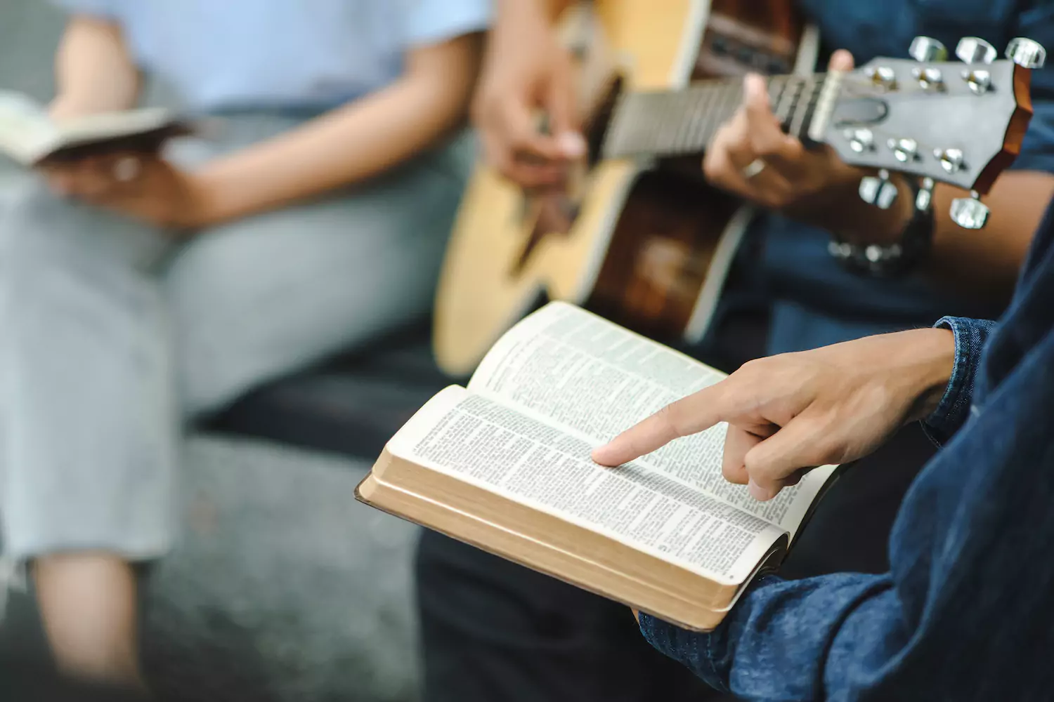 A person points at the Bible and follows along as another person strums a guitar in the background, leading a worship song.