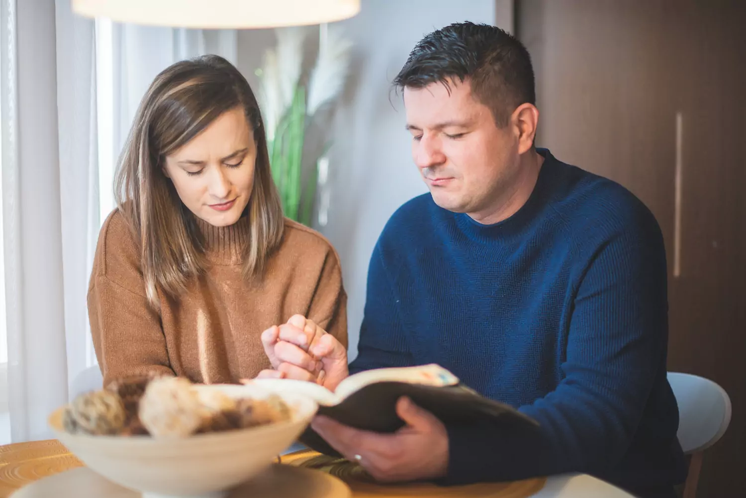 A husband and a wife sit at a table reading an open Bible with each other.