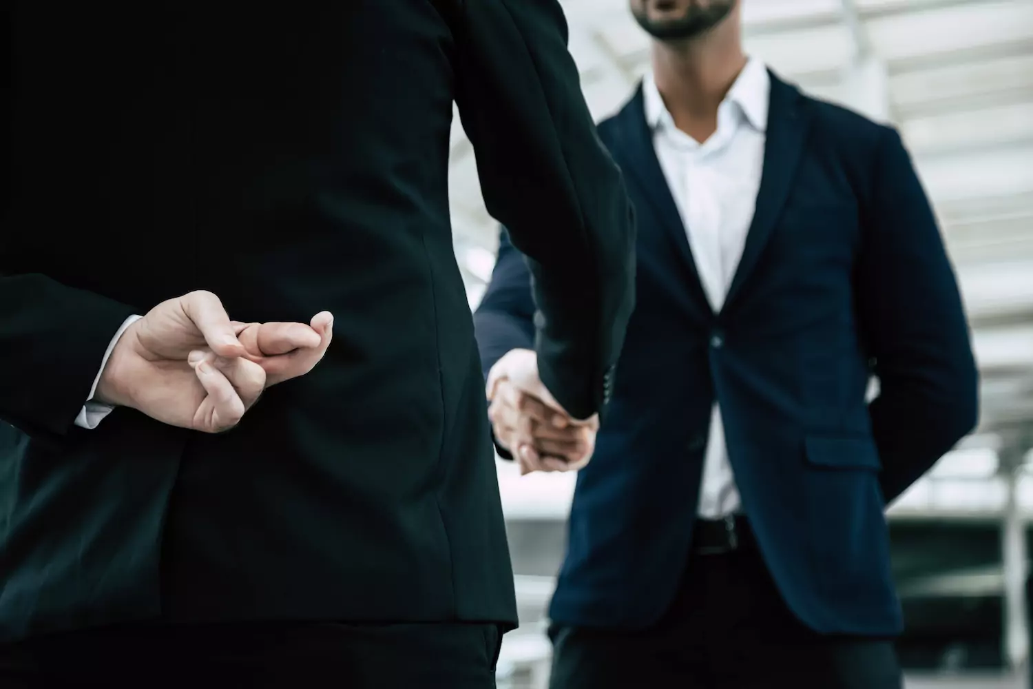 A man with his back to the camera crosses his fingers behind his back as he shakes the hand of another man, as if he intends to break his word and betray the other man.
