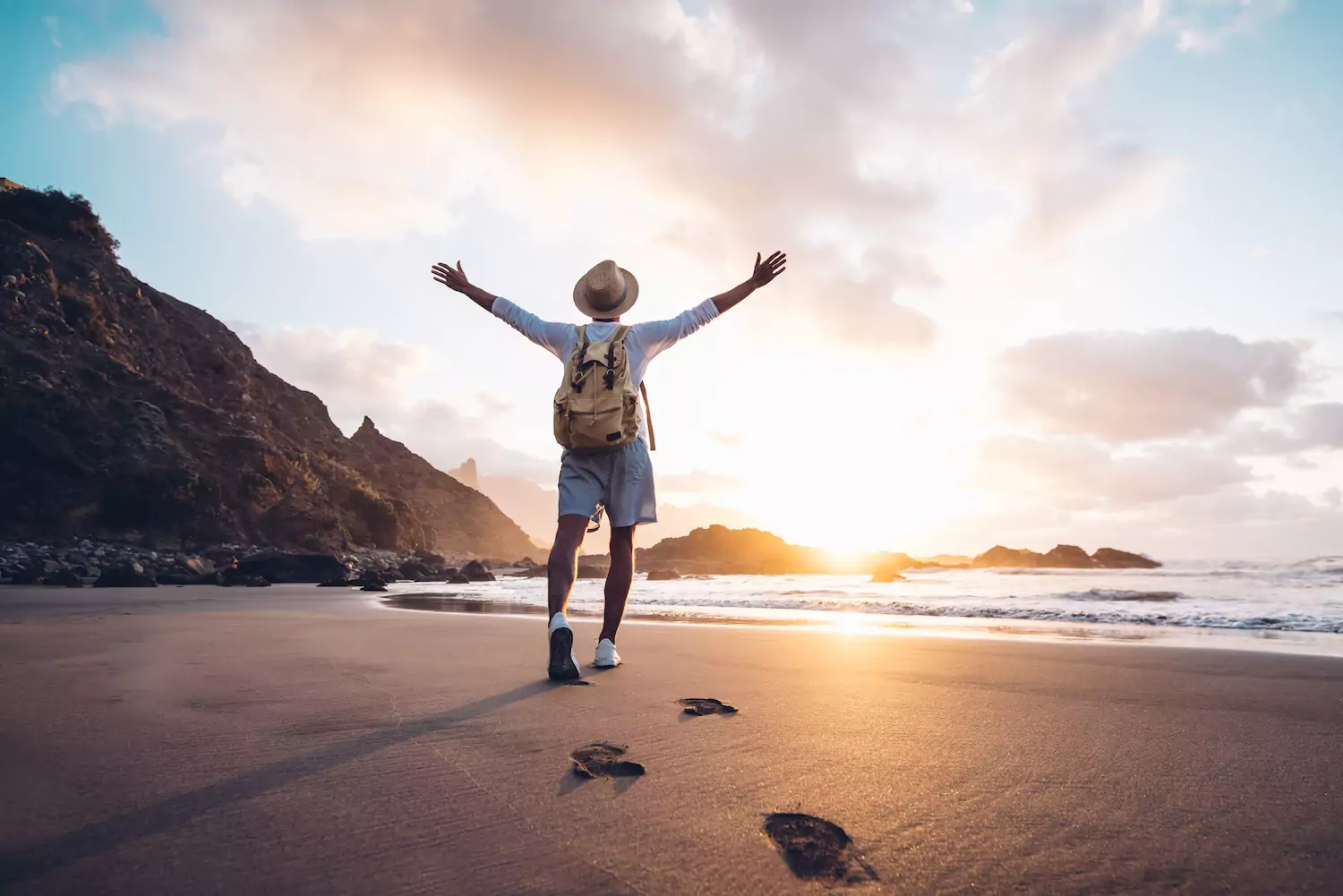 A man stands in a peaceful setting on a mountainous beach, arms outstretched, facing a soft sunset.