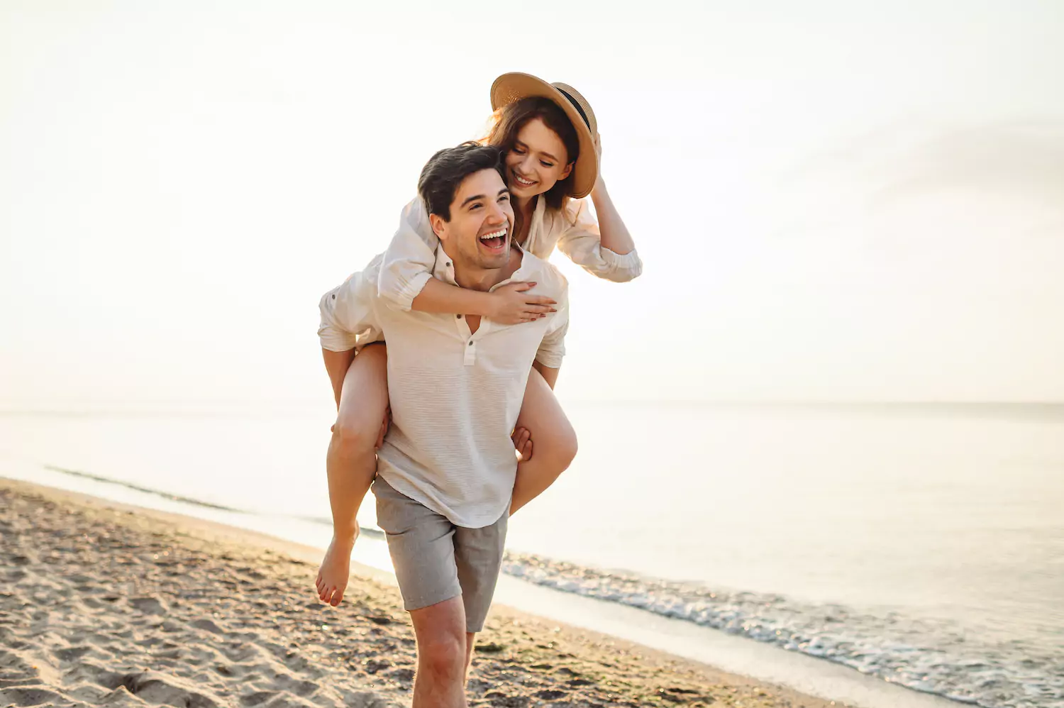 A man and an a woman walk on a beach at sunset. The man is giving the woman, who is wearing a sunhat, a piggyback ride.