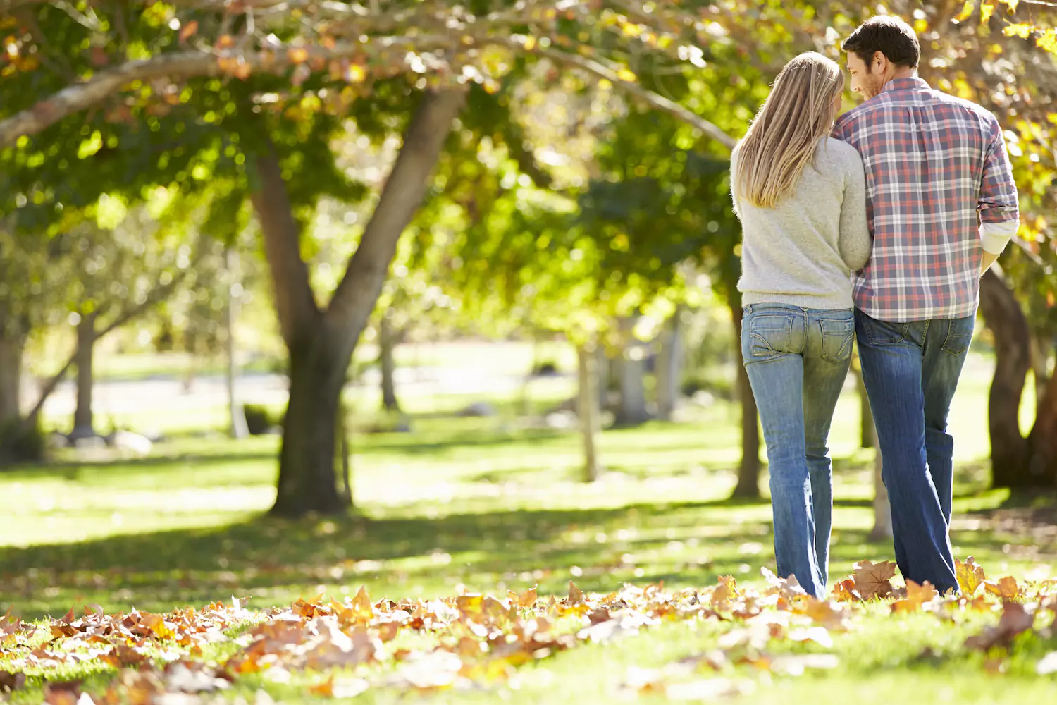 A husband and a wife walk arm-in-arm though a park with a canopy of trees and fallen leaves on the ground.