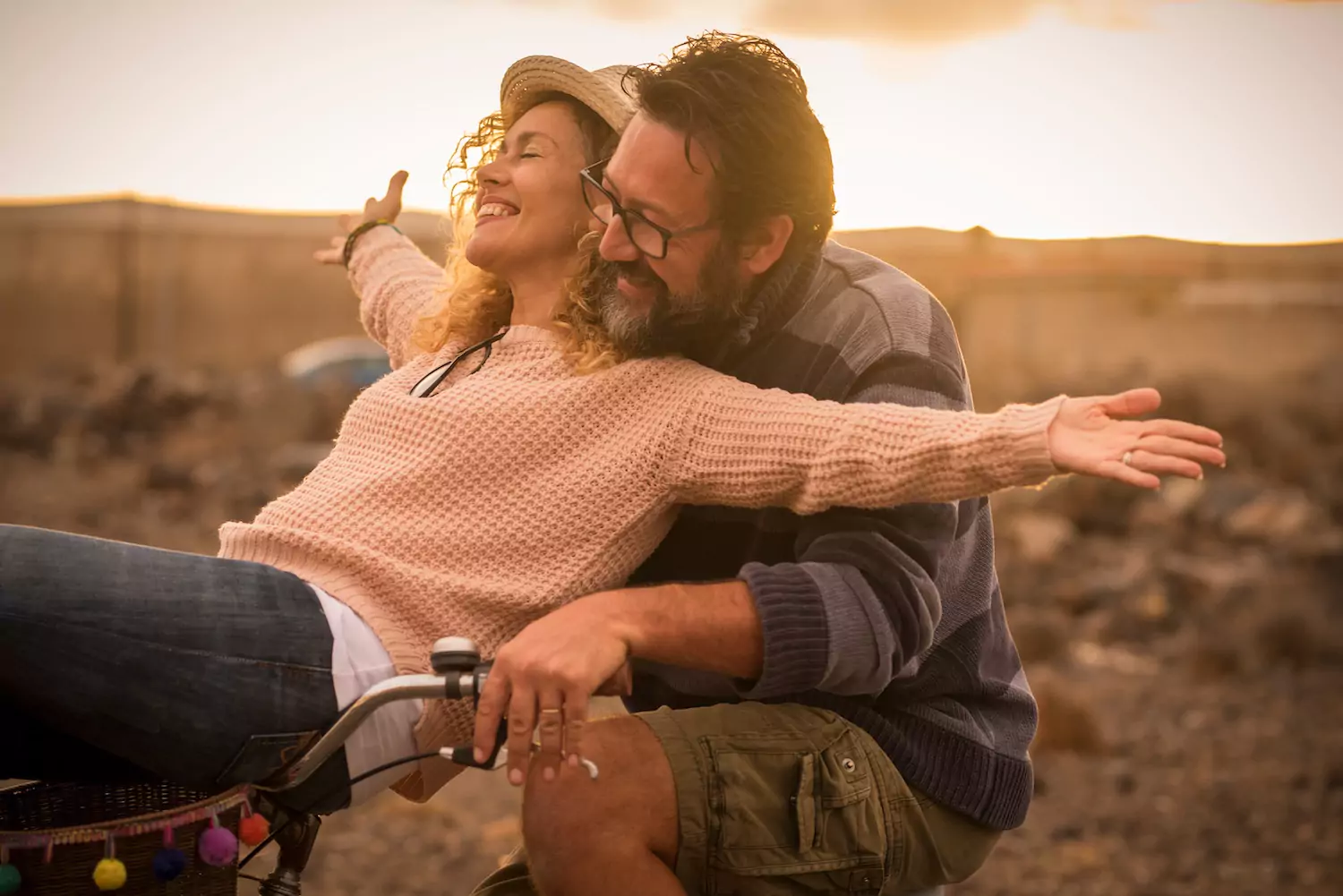 A photo of a man riding a bike at sunset, with his wife joyfully riding on his handlebars, leaning back on her husband with her arms outstretched in joy.