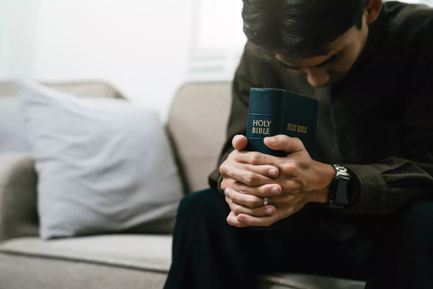 a man with dark hair sits with head bowed in prayer and hands folded around his Bible