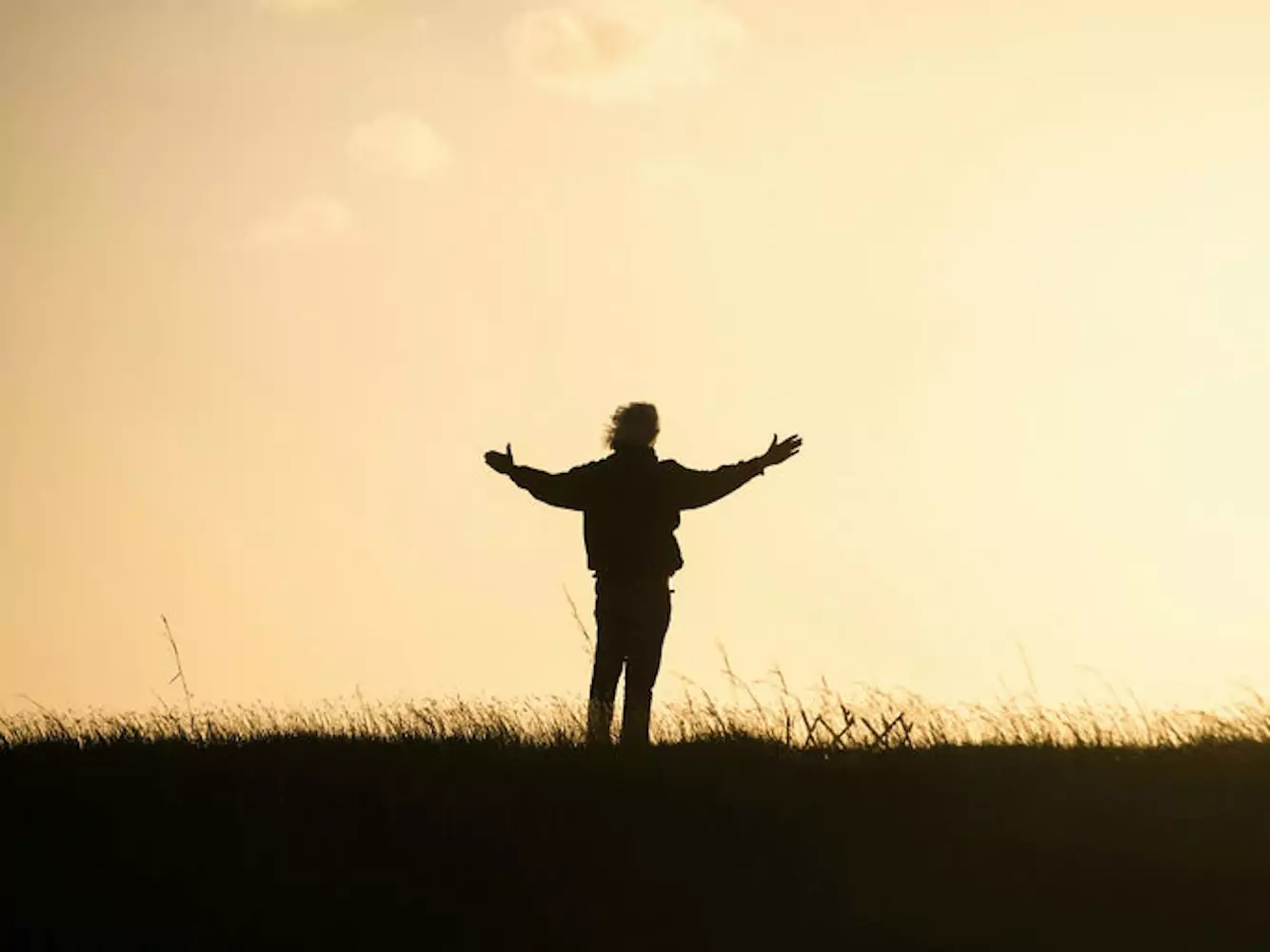 silhouette of a man with windblown hair and outstretched arms gazing at a golden sky