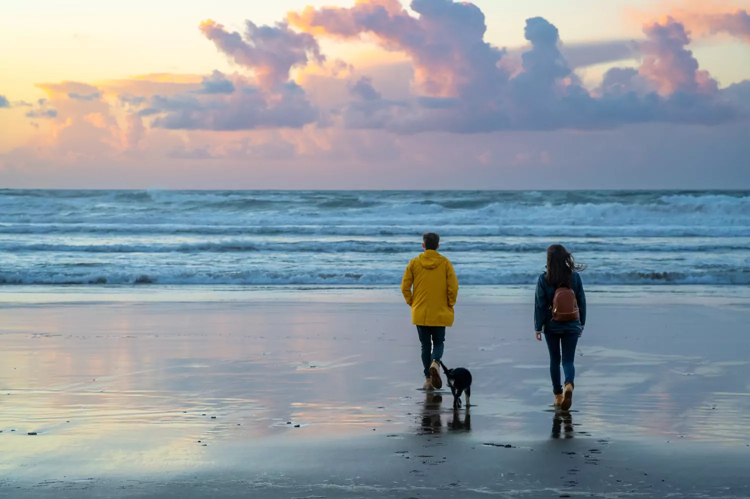 A pastor and his wife walk their dog on the beach beneath a cloudy, dusky sky.