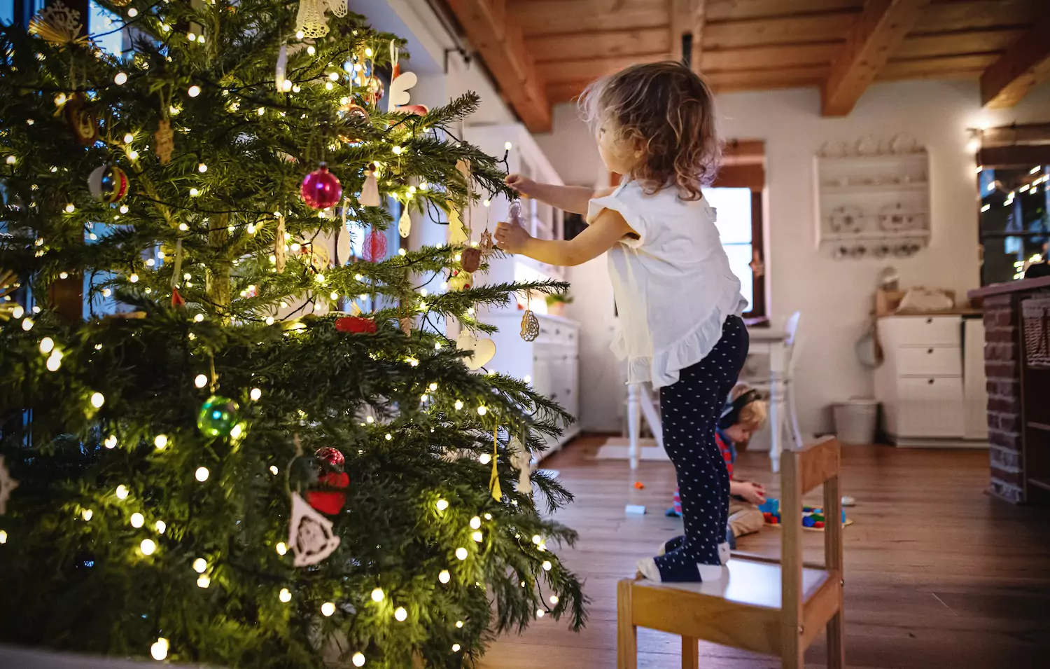 A little girl stands on a chair to hang an ornament on the family Christmas tree in celebration of advent.