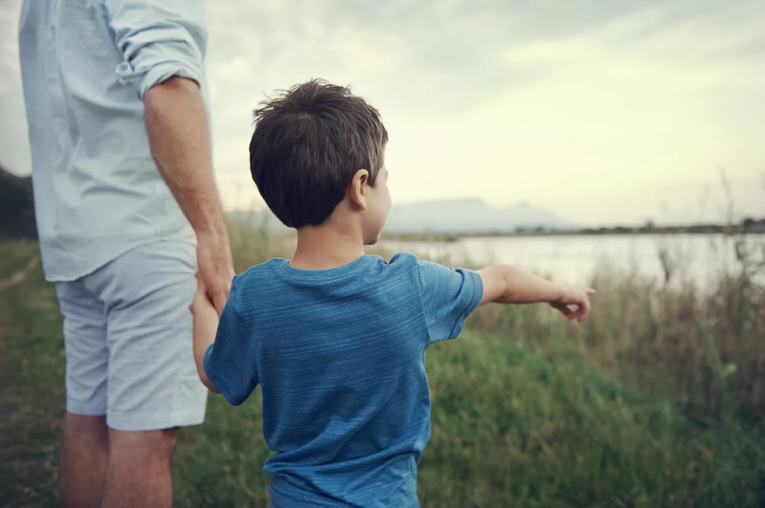 A pastor holds his son's hand as they stand in tall grass next to the lake.