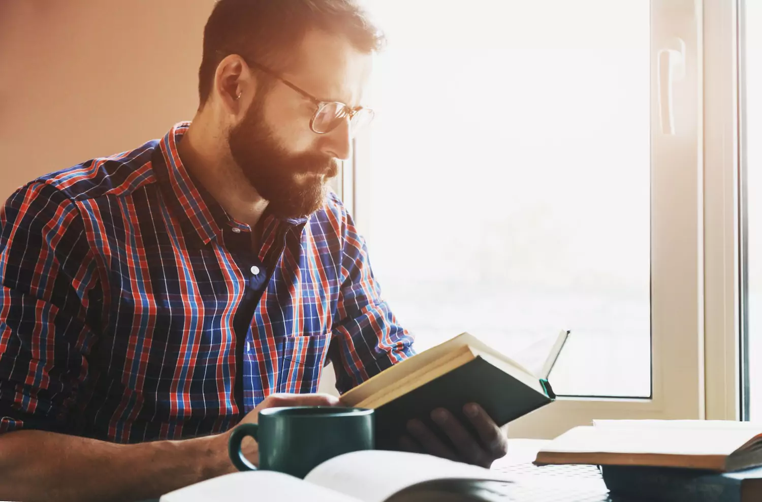 a pastor sits next to a window reading with his coffee mug next to him.